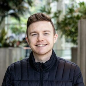 A person with short hair smiling while wearing a black quilted jacket. The background features indoor plants and blurred elements.