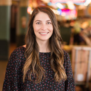 A woman with long brown hair, wearing a black patterned blouse, smiles while standing indoors. The background is softly blurred with colorful lights and a warm atmosphere.