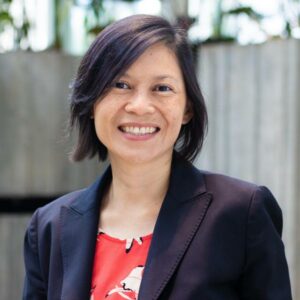 A person with shoulder-length dark hair and a friendly smile is wearing a black blazer over a red and white patterned top. They are standing indoors with a blurred background of vertical wooden panels and greenery.