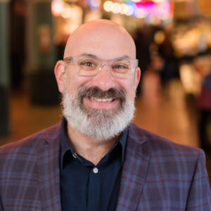 A bald man with a gray beard smiles while wearing glasses and a plaid jacket. The background is softly blurred, suggesting an indoor setting with warm lighting.
