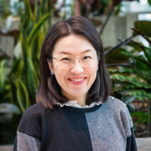 Smiling person with glasses and shoulder-length dark hair, wearing a black and gray sweater, stands in front of a lush, green plant background.