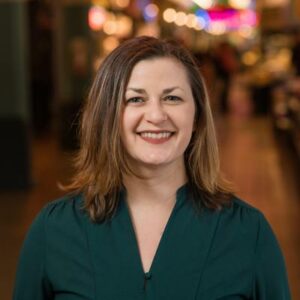 A woman with shoulder-length brown hair smiles warmly, wearing a dark green blouse. She stands in a softly lit indoor setting with blurred lights in the background.