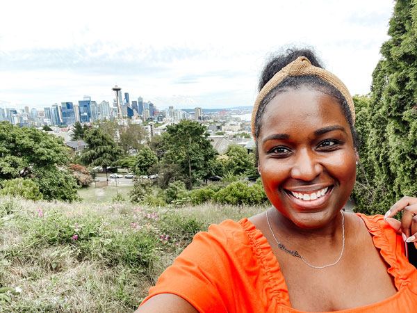 A woman in an orange top smiles for a selfie in a park overlooking a city skyline with a prominent tower. The foreground is filled with greenery and trees. The sky is partly cloudy.
