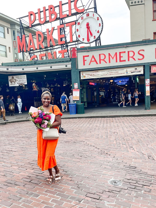 A person in an orange dress stands smiling, holding a bouquet of flowers in front of the entrance to a public market. The market has a large clock and signs that read "Public Market Center" and "Farmers Market." The ground is cobblestone.