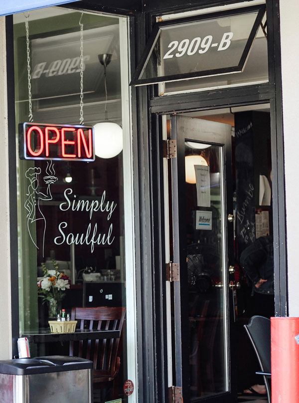 The image shows the entrance of a restaurant named "Simply Soulful" with a neon "Open" sign. The door is partially open, revealing a glimpse of tables and flowers inside. The address "2909-B" is displayed above the entrance.