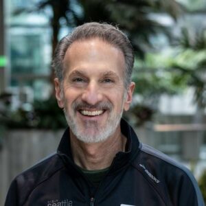 A smiling man with short gray hair and a beard stands in a bright indoor setting with plants in the background. He is wearing a dark jacket with a small logo on the chest.