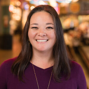 A woman with long dark hair smiles warmly. She is wearing a purple top and a necklace. The background is softly blurred, featuring indoor lighting and indistinct shapes that suggest a market or store setting.