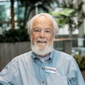 Elderly person with a white beard and light blue shirt, wearing a name tag, smiles at the camera. The background shows greenery and a softly lit indoor area.