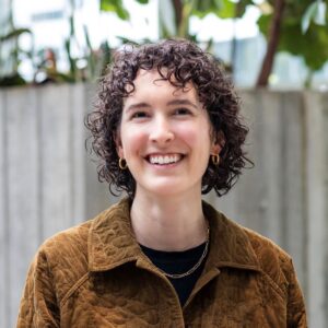 A person with curly hair smiles warmly. They wear a brown jacket, gold hoop earrings, and a gold chain necklace. The background features blurred greenery and concrete.