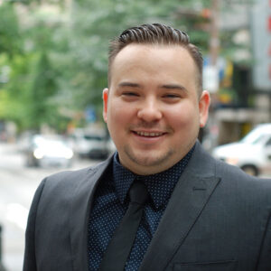 A person in a suit and tie smiles at the camera while standing on a city street. The background features blurred greenery and buildings.