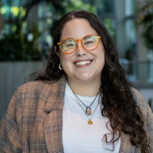 A smiling person with long curly hair and orange glasses is wearing a plaid blazer and white shirt. They are in a bright indoor space with plants in the background.