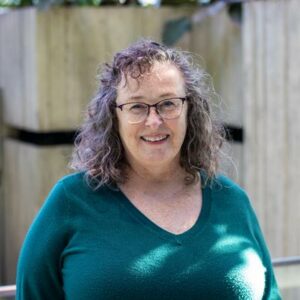 A woman with curly hair and glasses is smiling at the camera. She is wearing a teal sweater and standing in front of a wooden structure with plants.