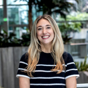 A smiling person with long blonde hair wearing a black shirt with white stripes stands indoors. The background features green plants and large windows, creating a bright and natural atmosphere.